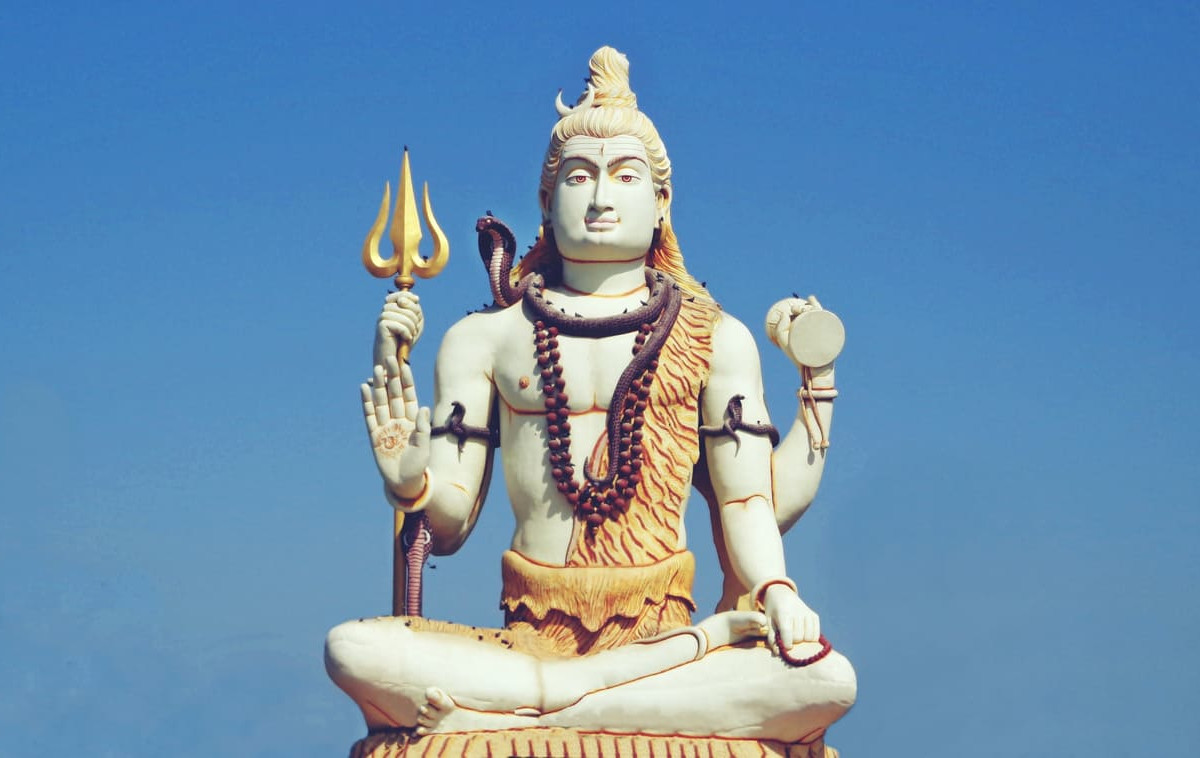 A towering statue of Lord Shiva in a meditative pose, holding a trident and drum, set against a clear blue sky.