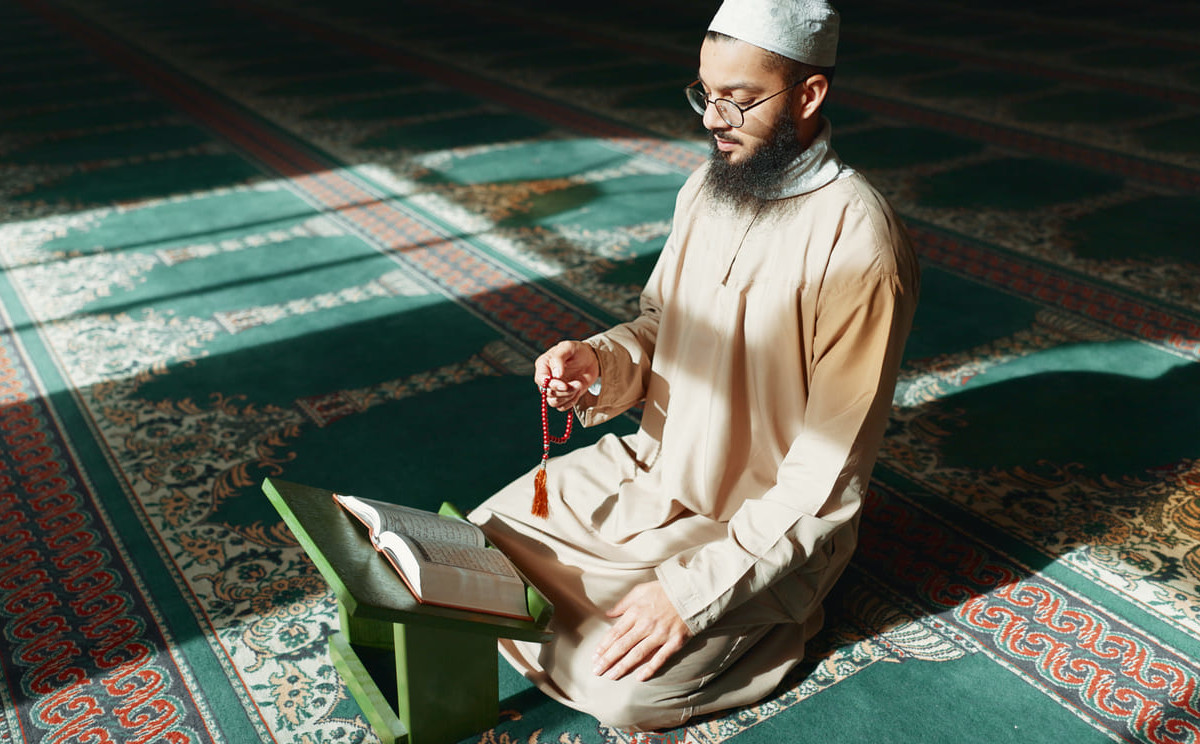 A Muslim man sits in a sunlit mosque, holding prayer beads and reading the Quran in quiet reflection.