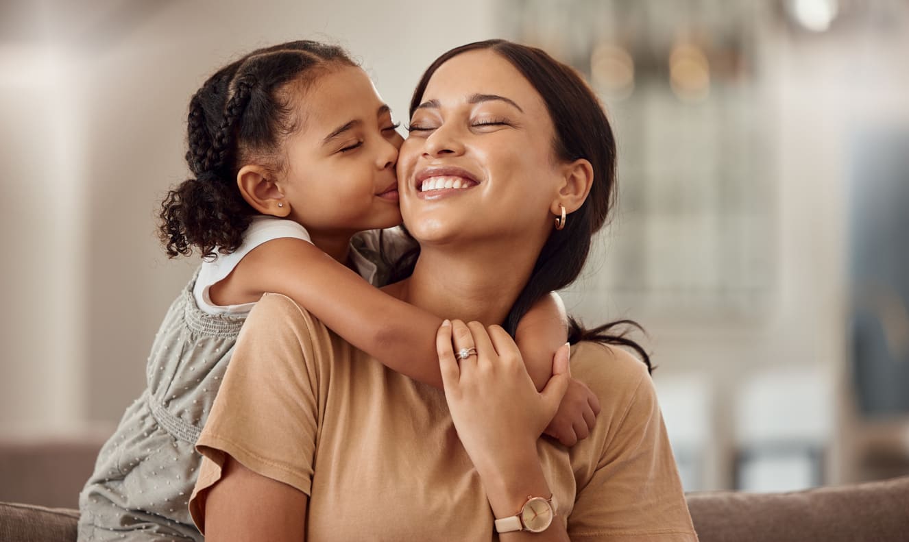 A young girl lovingly hugs and kisses her smiling mother, expressing affection in a warm and heartfelt moment.