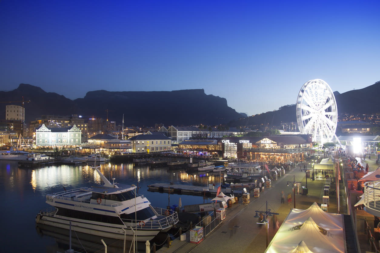 Cape Town's V&A Waterfront at dusk, featuring a lit-up ferris wheel, lively crowds, and boats docked along the harbor against the backdrop of Table Mountain.