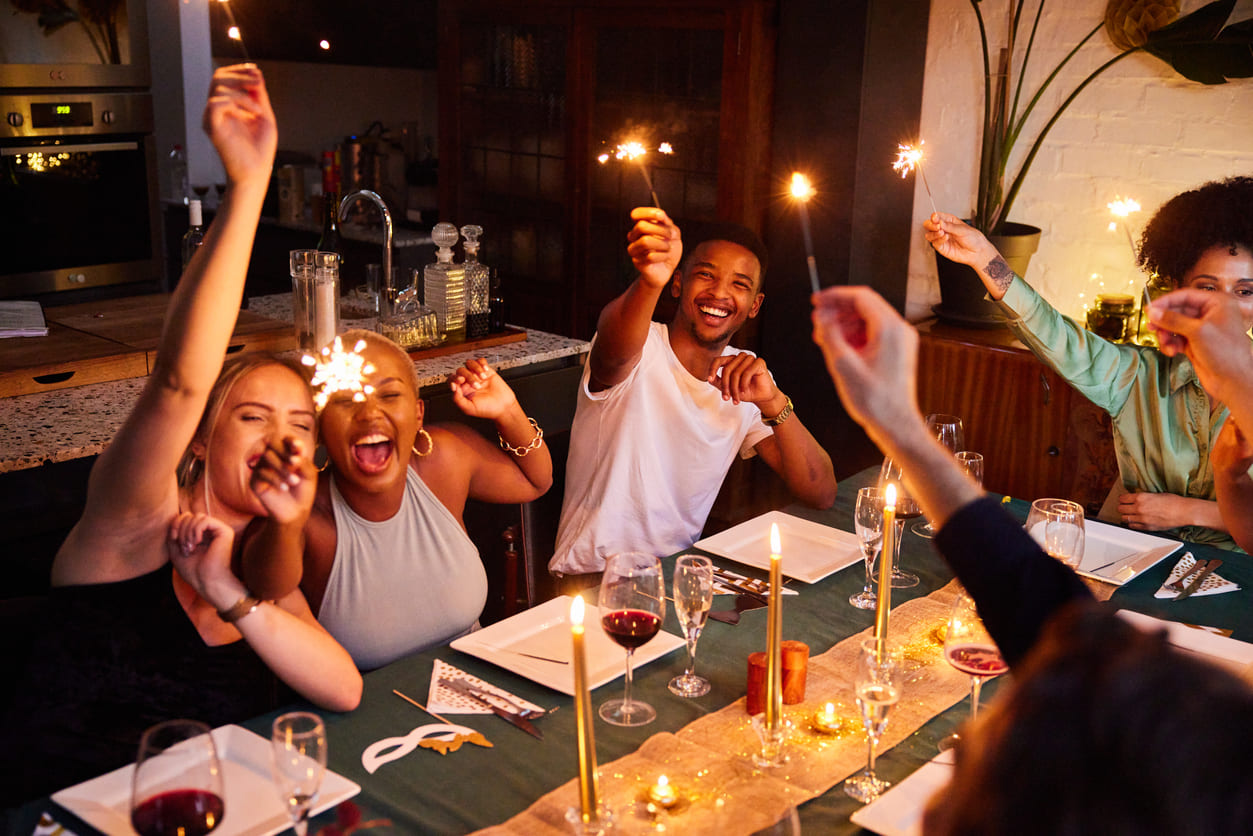 A lively group of friends gathered around a festive dinner table, holding sparklers and celebrating with smiles and laughter.