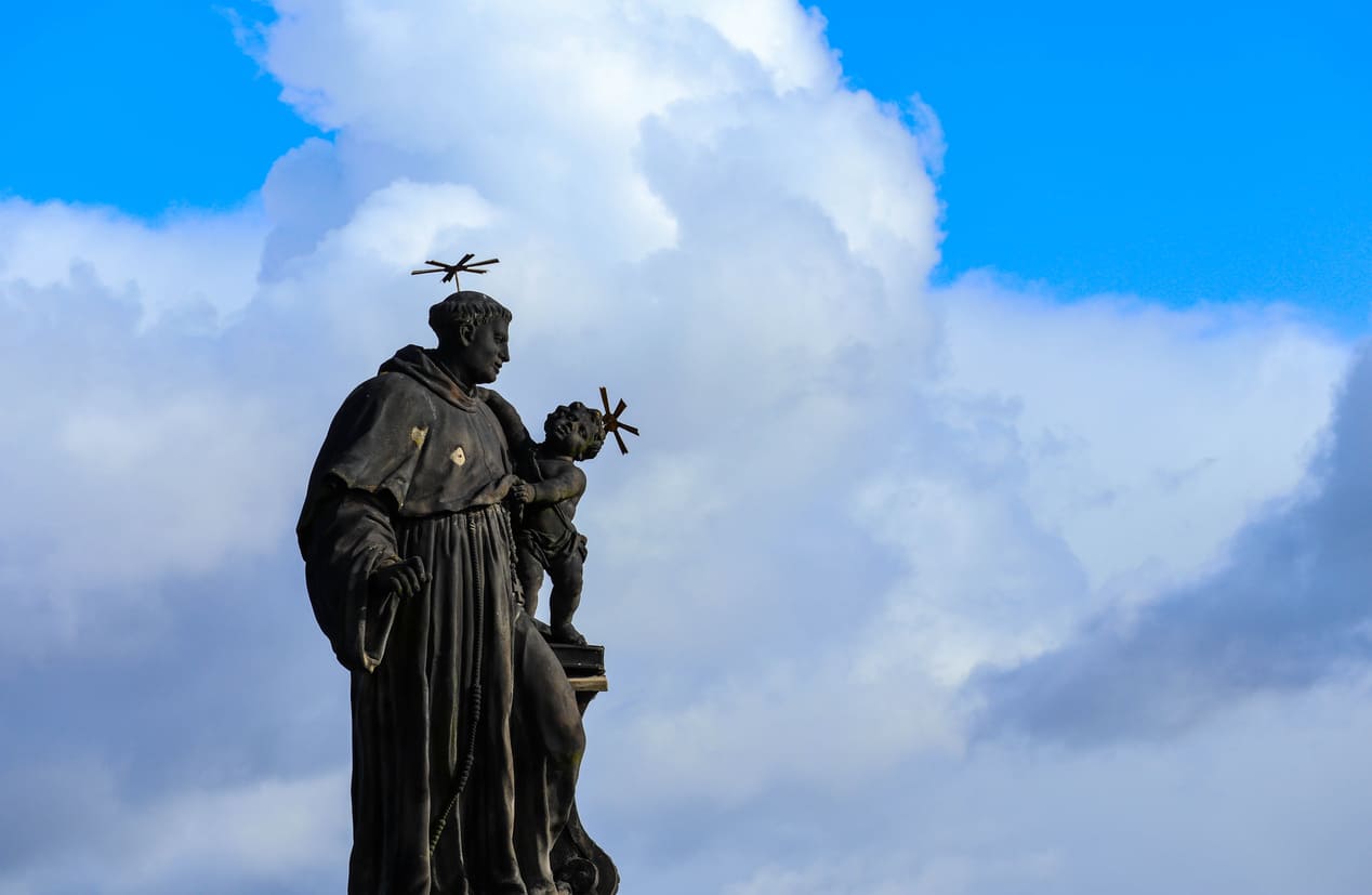 A statue of Saint Anthony of Padua holding the Child Jesus, adorned with halos, set against a backdrop of a bright blue sky and clouds.