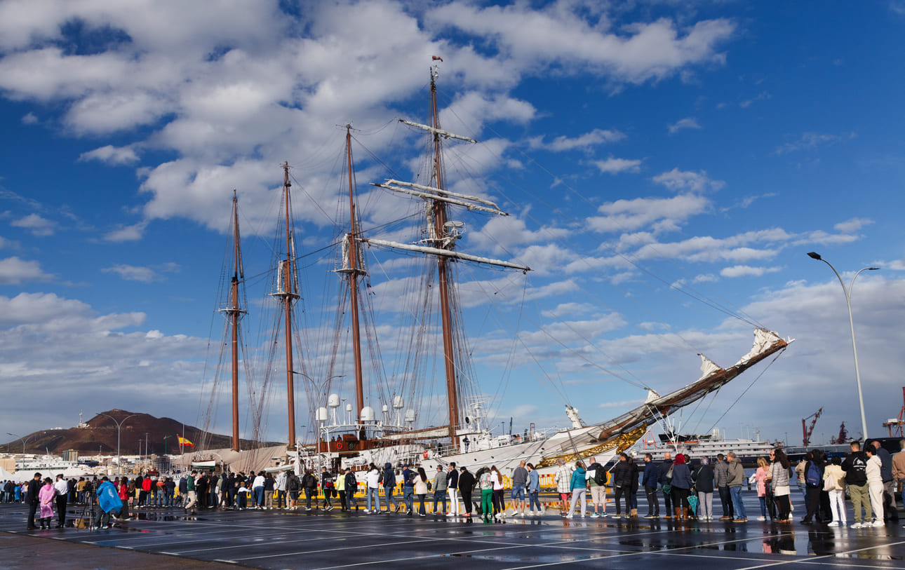 The Spanish training ship Juan Sebastián de Elcano is moored at the Naval Base, with visitors lining up to explore its historic decks under a bright blue sky.
