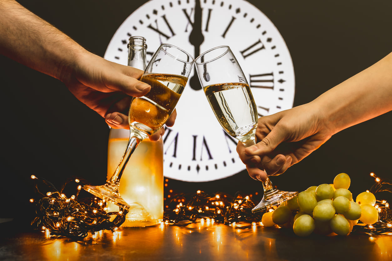 A festive New Year's Eve scene featuring a midnight toast with champagne, a clock striking twelve, twinkling lights, and the traditional twelve grapes for good luck.