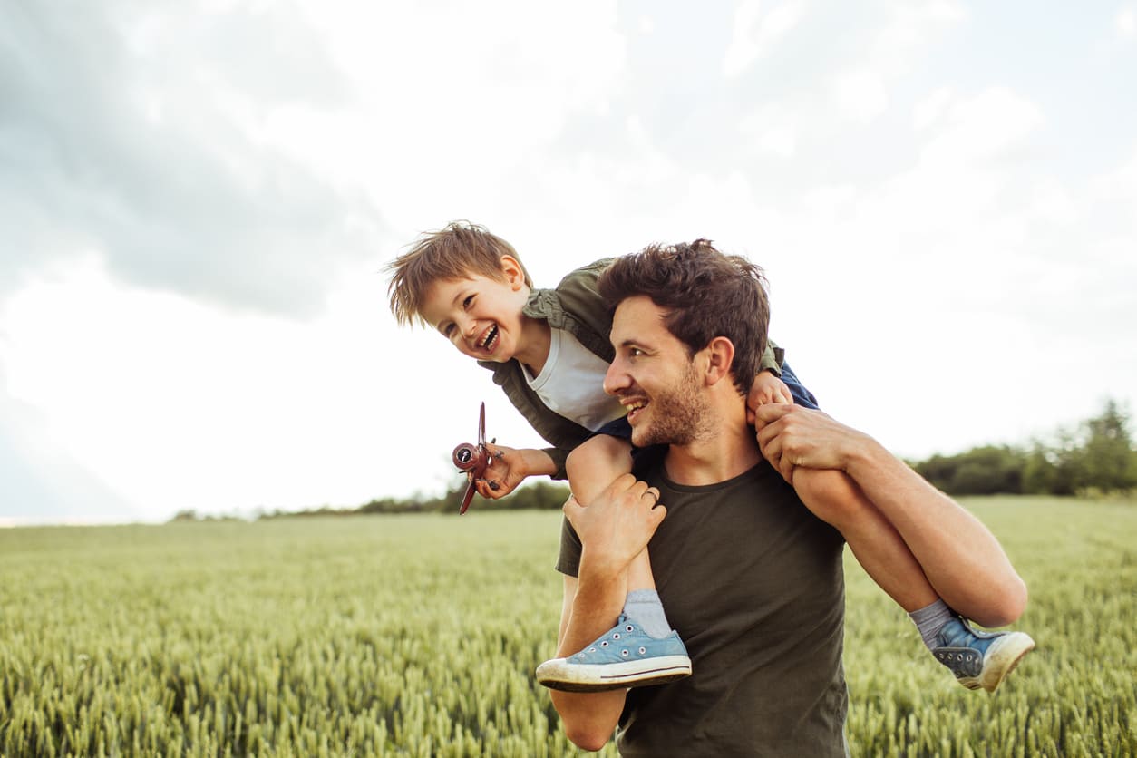 A smiling father carries his son on his shoulders in a green field, capturing a playful and heartwarming moment on Father's Day.