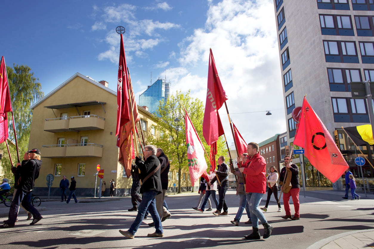 A group of people walk through a city street holding red flags, participating in a peaceful Labour Day march under a bright spring sky.