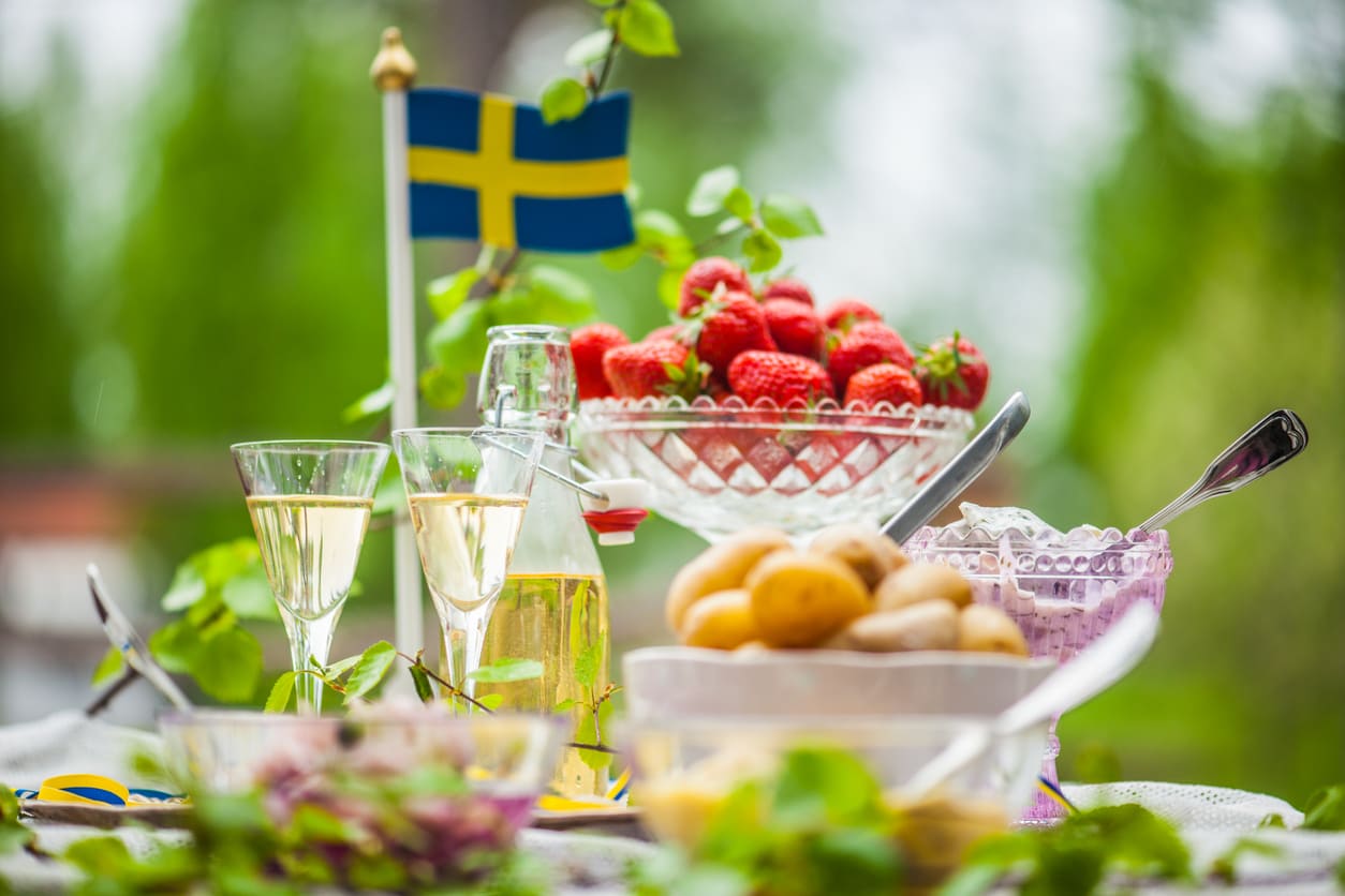 A vibrant outdoor Midsummer table with classic Swedish dishes, fresh strawberries, snaps, and a Swedish flag surrounded by summer greenery.
