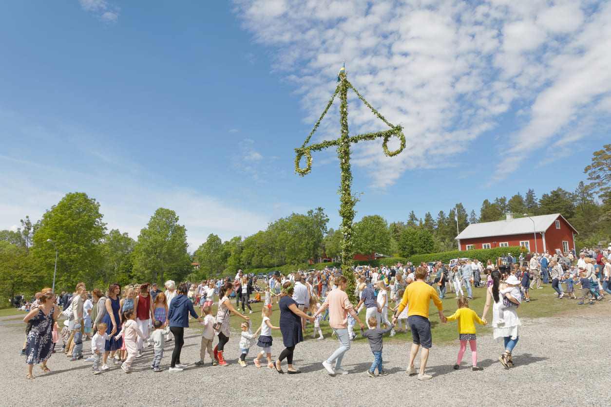 A lively outdoor scene with people of all ages dancing around a tall, flower-decorated maypole under a bright summer sky.