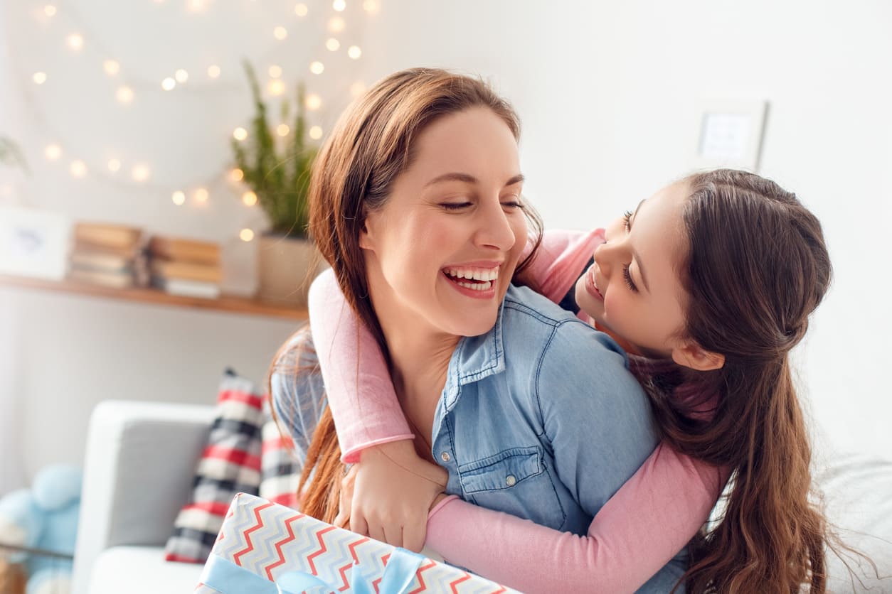 A mother and daughter share a happy embrace at home, surrounded by soft lighting and a wrapped gift, reflecting the warmth of Mother's Day.