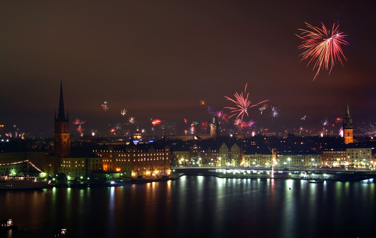 A festive night view of Stockholm with fireworks bursting over the city skyline and reflections shimmering on the water.