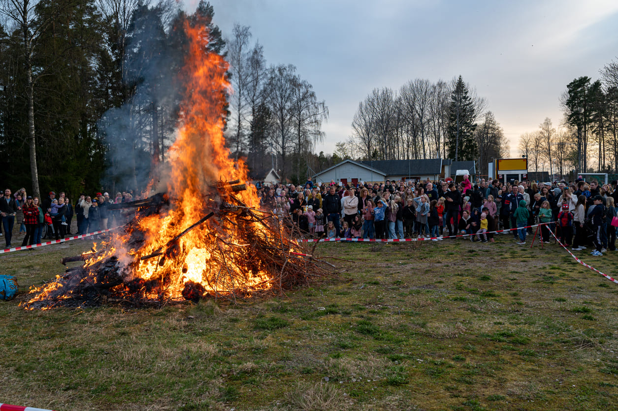A crowd of people, including families and children, stands around a large bonfire in an open field, marking the traditional Walpurgis Night celebration.