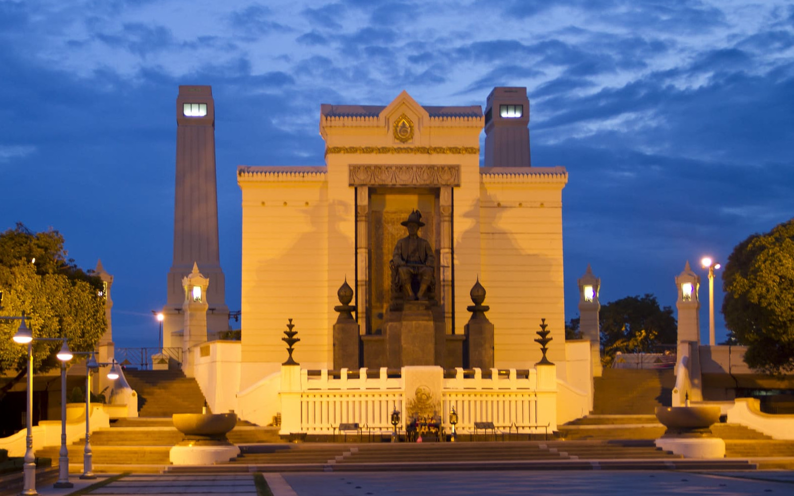 A well-lit statue of King Rama I at dusk, framed by grand architecture and a deep blue sky in Bangkok.