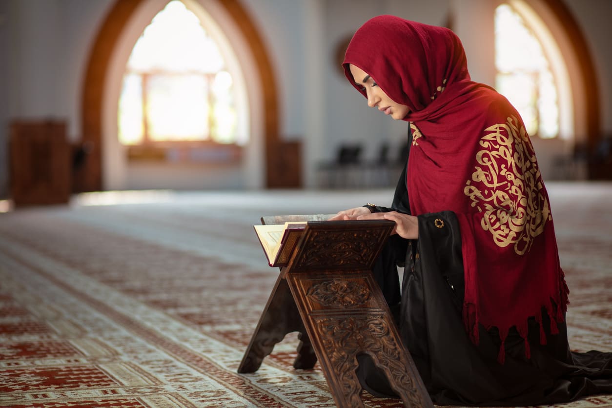 A woman sits in quiet devotion, reading from a holy book inside a beautifully lit mosque.